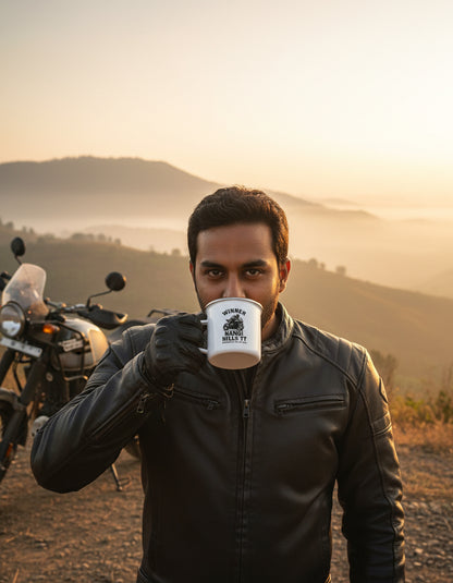 Man drinking from white mug with black text and graphics near a hill with a bike